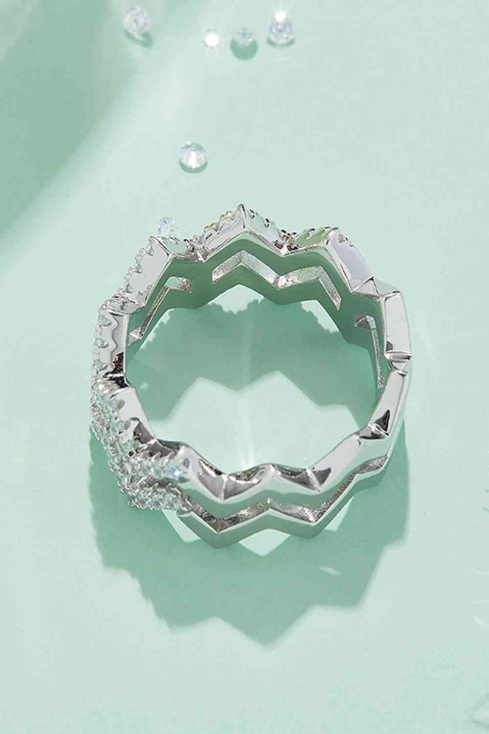 A silver Adored Moissanite Zigzag Stacking Ring from Marianela's Exclusive Shop, LLC with a unique design rests on a light blue surface. The ring features a row of small, sparkling gemstones along part of its exterior, adding a touch of elegance to its geometric shape. Small scattered gemstones are seen in the background, highlighting this fashion-forward jewelry piece.