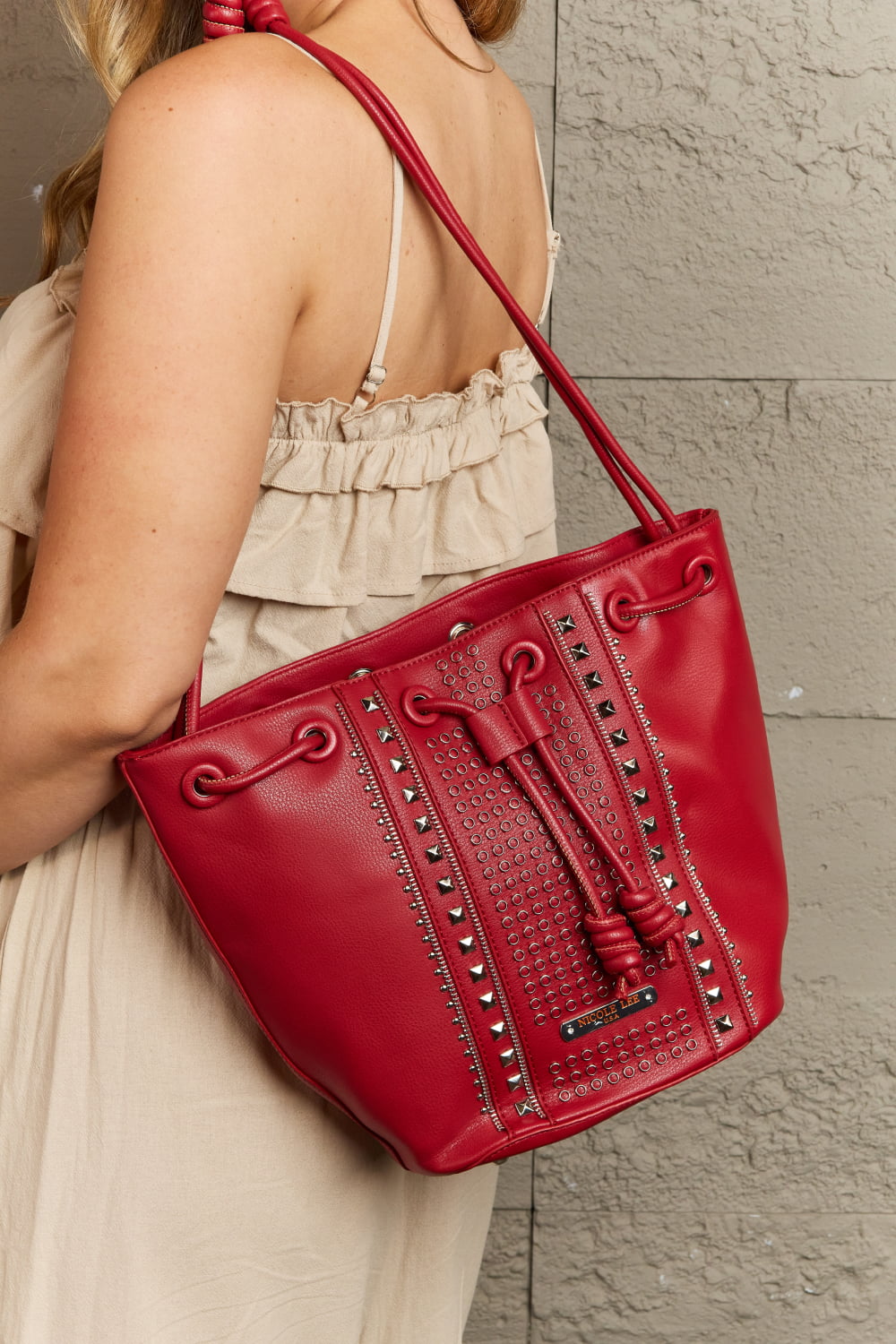 A person with light wavy hair, wearing a beige sleeveless dress, is shown from the back. They carry a red Nicole Lee USA Amy Studded Bucket Bag from Marianela's Exclusive Shop, LLC over their shoulder. The handbag features pebbled vegan leather adorned with stud detailing and a secure drawstring closure. The background is a gray textured wall.