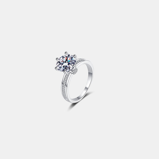 A 3 Carat Moissanite 925 Sterling Silver Engagement Ring from Marianela's Exclusive Shop, LLC is adorned with smaller diamonds on its band, placed on a plain white background. The Moissanite sparkles, reflecting light and showcasing an intricate and elegant design.