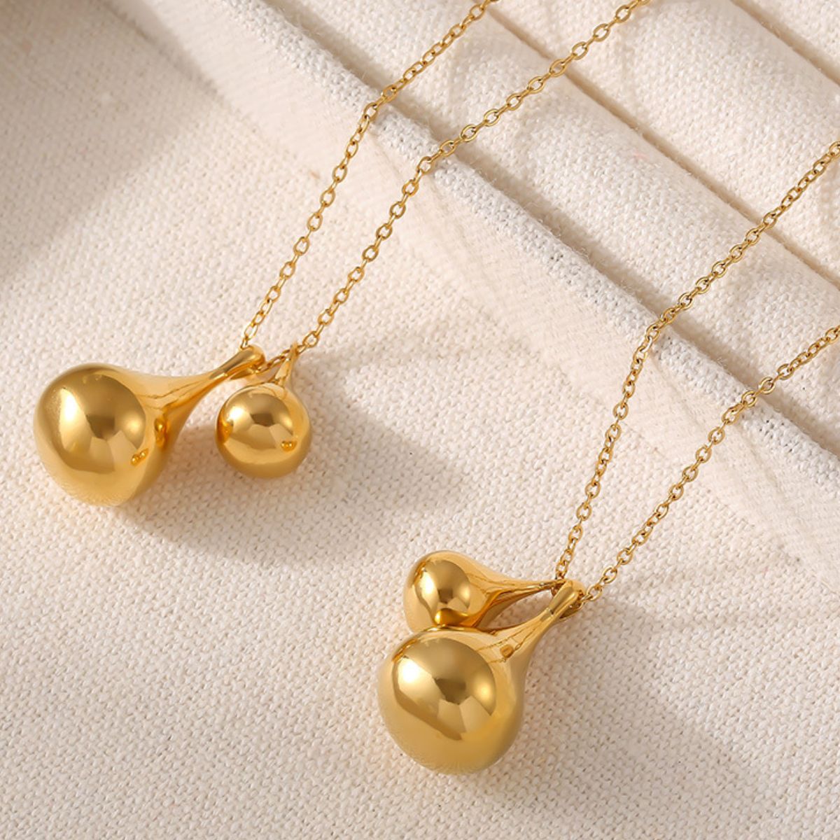 Displayed on light beige fabric, the Elysian Gold Water Drop Necklaces from Marianela's Exclusive Shop feature elegant 18K gold-plated designs that resemble smooth, rounded water drop pendants. Delicate chains enhance the refined appearance of these exquisite pieces.