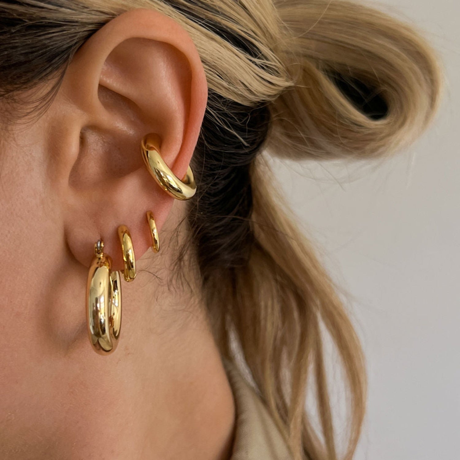 Close-up of a person's ear adorned with four Stainless Steel Letter C Shape Clip On Earrings from Marianela's Exclusive Shop, LLC of decreasing size. The person has light hair tied back, and the earrings are positioned from the lobe up through the cartilage. The background is a plain, off-white color.