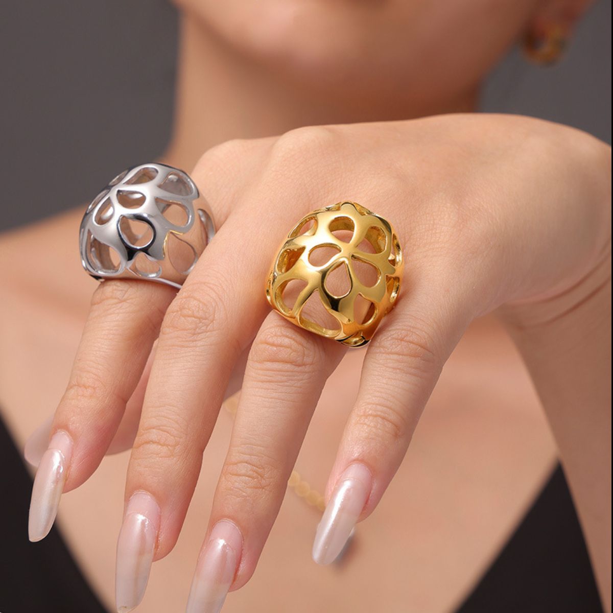 A person wearing two large, floral-pattern rings, one an 18K gold-plated piece and the other the Gold-Cutout Elegance Ring from Marianela's Exclusive Shop. Their nails are long and polished, embodying modern minimalism against a neutral background.