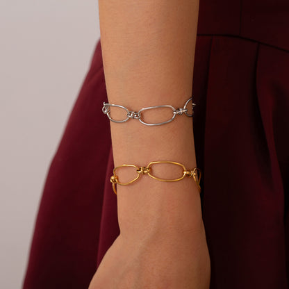 A person flaunts two chain-link bracelets on their wrist—a sleek Titanium Steel Chain Bracelet paired with Marianela's Exclusive Shop's Gold Hollow Chain Bracelet—set against the rich backdrop of a burgundy garment, highlighting a modern link design.