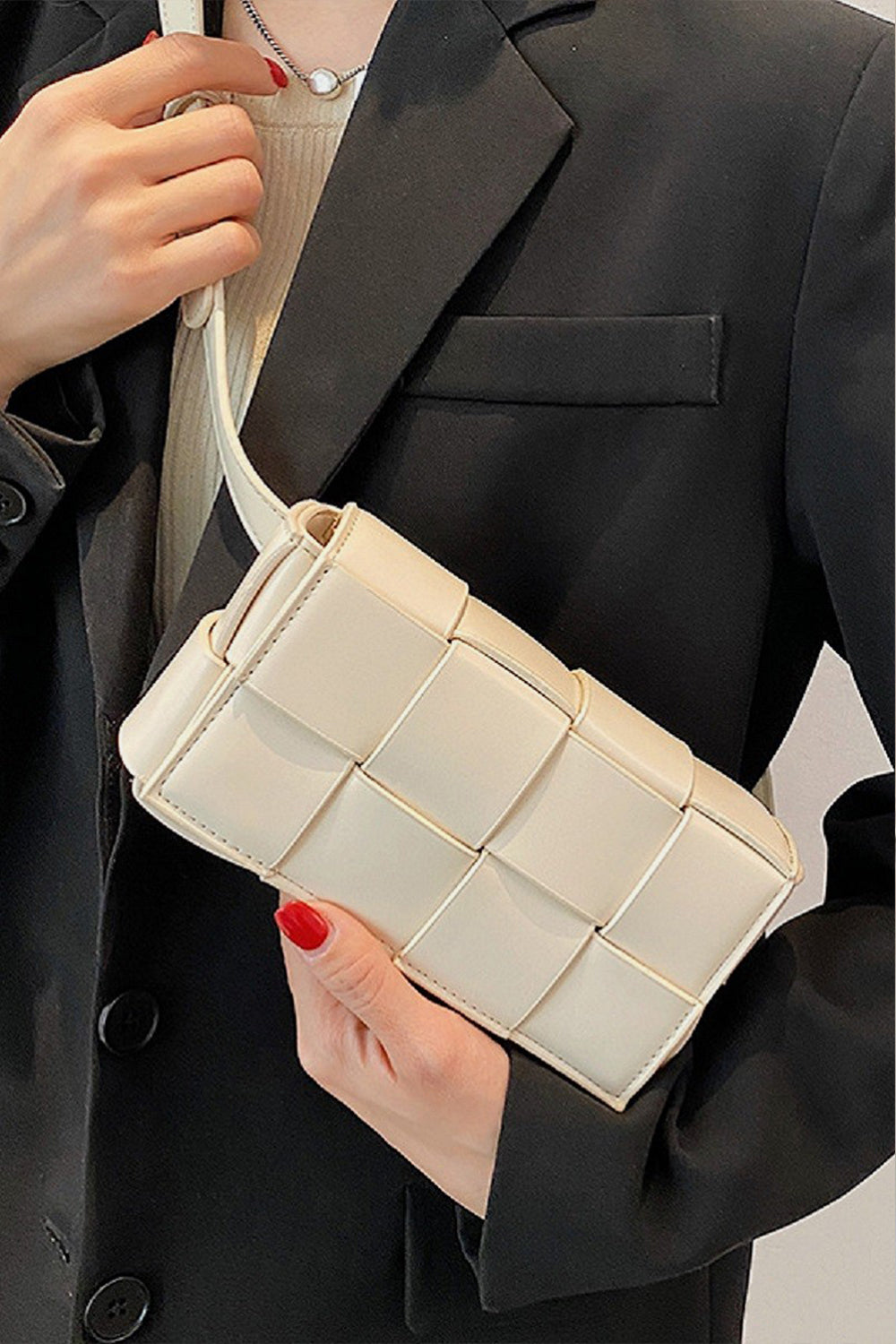 A person wearing a black blazer holds a cream-colored Zenana Vegan Leather Woven Crossbody Bag from Marianela's Exclusive Shop, LLC with one hand. The person has a red manicure and is wearing a beige top underneath the blazer. A delicate necklace is partially visible above the top, adding an eco-friendly touch to their chic ensemble.