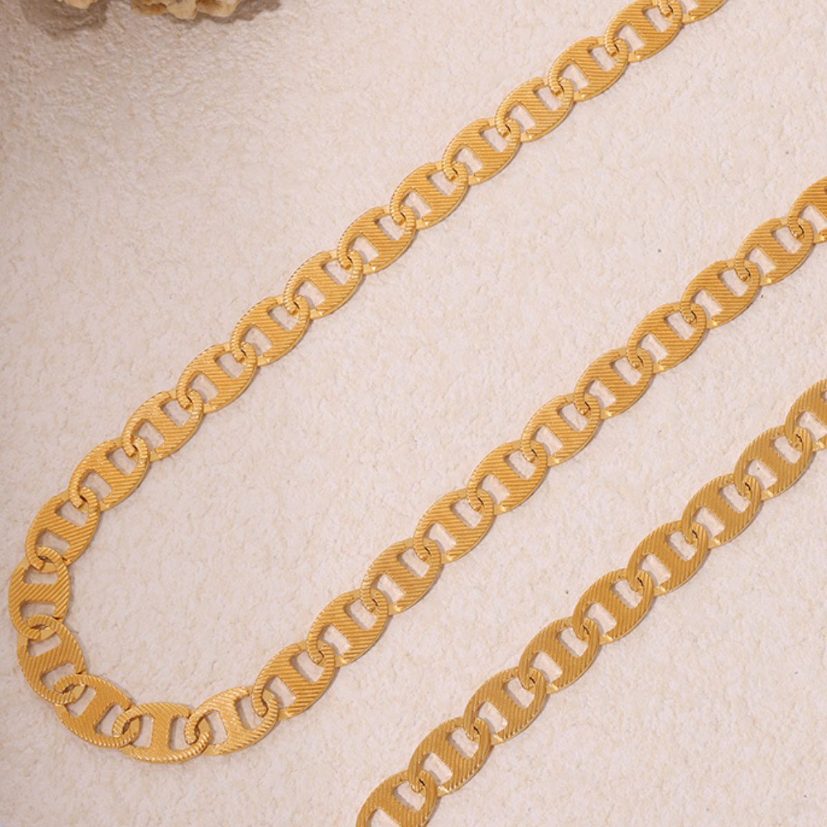 The Luxe Gold Chain Necklace from Marianela's Exclusive Shop features a minimalist design with exquisite 18K gold-plated links. Each oval segment is delicately arranged on a textured beige surface, highlighting intricate detailing that exudes luxury and elegance.