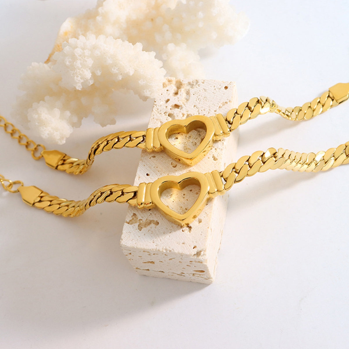 Two Titanium Steel Cutout Heart Bracelets from Marianela's Exclusive Shop, LLC are displayed on a light-colored textured stone, with a piece of white coral in the background. The shiny link chains and cutout heart accents create a stylish, romantic design.