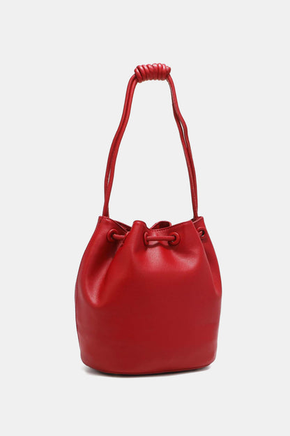 The Nicole Lee USA Amy Studded Bucket Bag, available at Marianela's Exclusive Shop, LLC, is a vibrant red bucket bag crafted from pebbled vegan leather. It features a secure drawstring closure at the top and a single strap with intricate knotted details, perfect for shoulder or hand carrying. Stud detailing adds an extra touch of style against the plain white background.