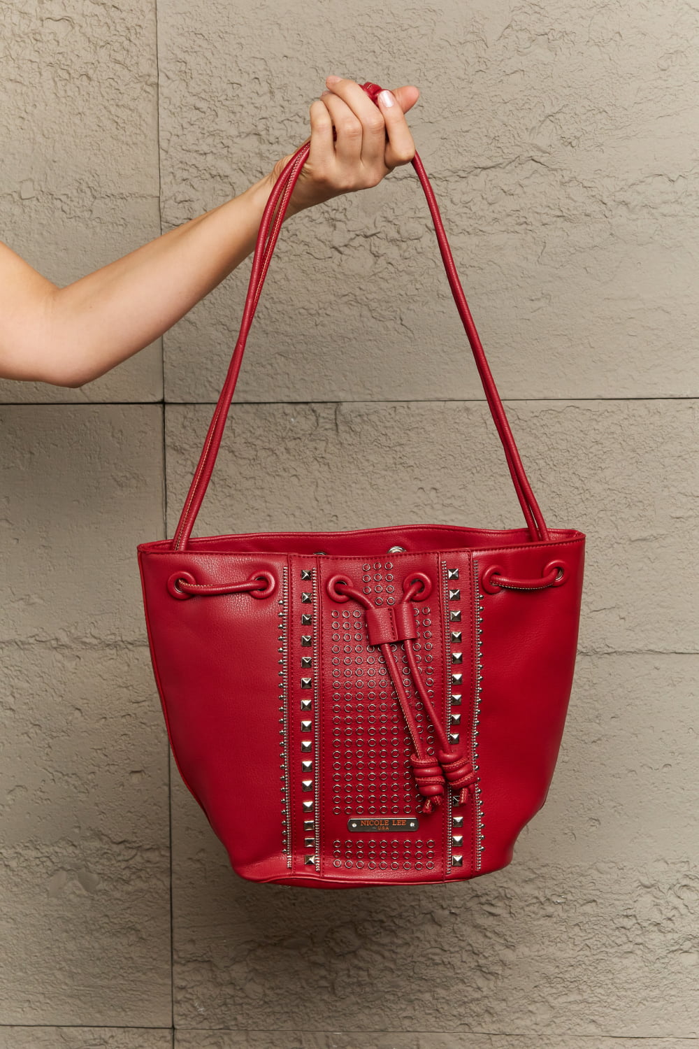 An individual is holding a Nicole Lee USA Amy Studded Bucket Bag in red, crafted from pebbled vegan leather with two handles, against a textured beige wall. The bag, available at Marianela's Exclusive Shop, LLC, boasts a secure drawstring closure and decorative metal studs arranged in vertical lines on the front.