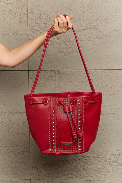 An individual is holding a Nicole Lee USA Amy Studded Bucket Bag in red, crafted from pebbled vegan leather with two handles, against a textured beige wall. The bag, available at Marianela's Exclusive Shop, LLC, boasts a secure drawstring closure and decorative metal studs arranged in vertical lines on the front.