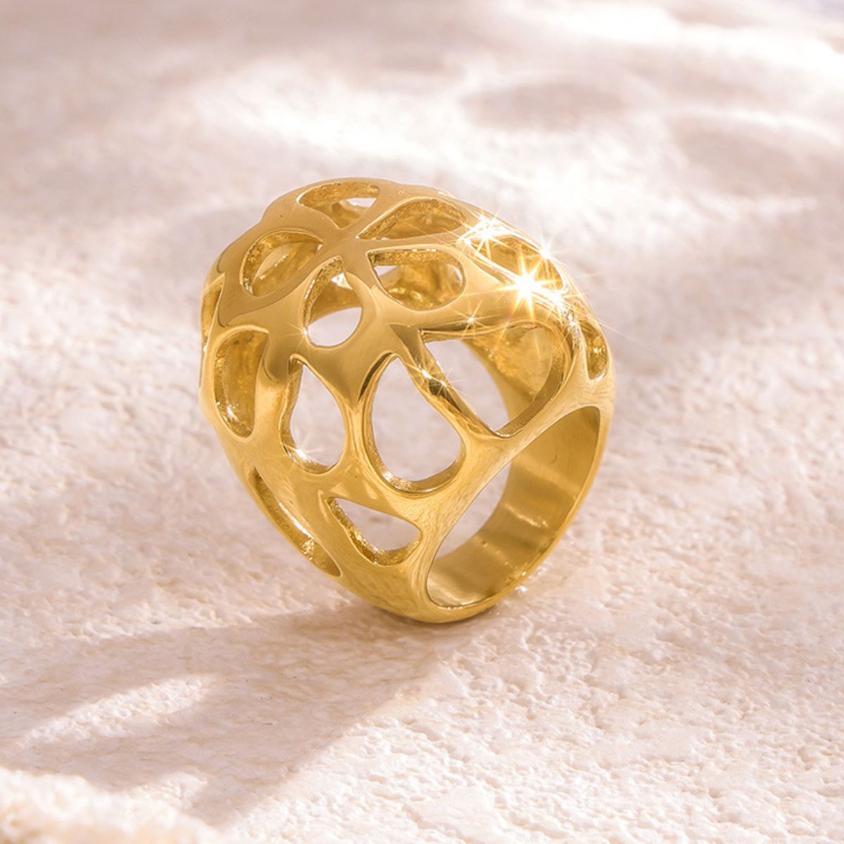 The Gold-Cutout Elegance Ring from Marianela's Exclusive Shop features an intricate openwork design in 18K gold plating. It beautifully reflects light and casts subtle shadows on a textured, light-colored background, embodying modern minimalism with its sophisticated allure.