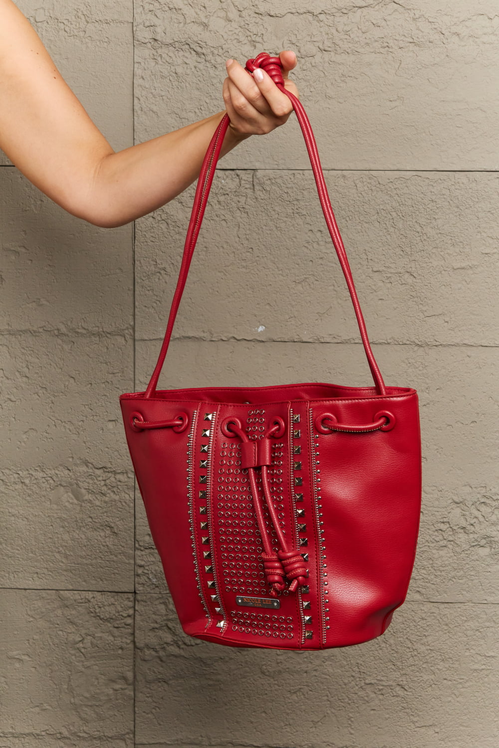 A person holds the Nicole Lee USA Amy Studded Bucket Bag, showcasing its secure drawstring closure. The bag, sold by Marianela's Exclusive Shop, LLC, is crafted from pebbled vegan leather and features decorative studs along with a textured pattern on the front. The background is a textured gray wall.