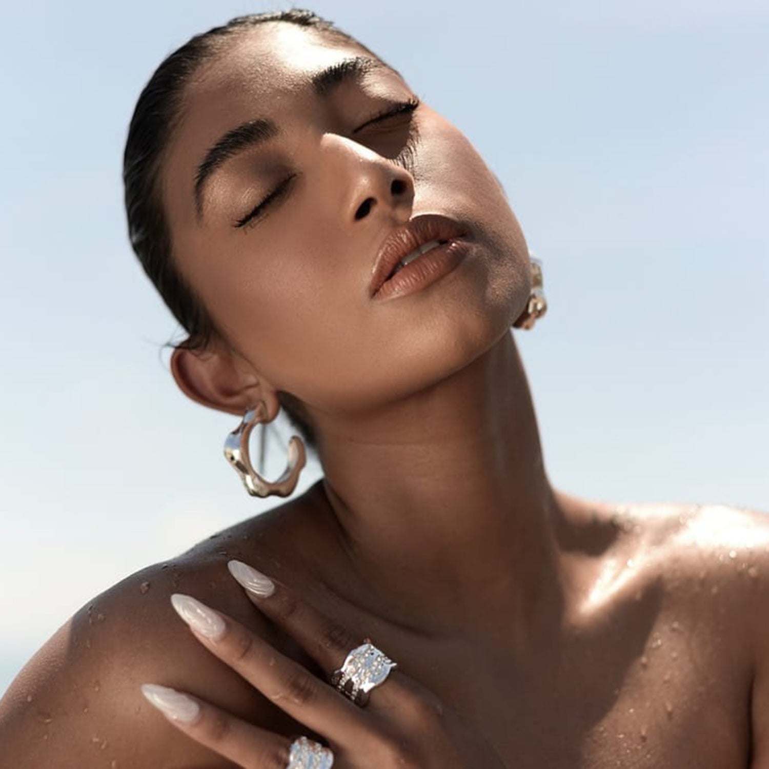 A woman with a sun-kissed complexion poses with her eyes closed and face lifted towards the sky. She wears large, stainless steel C-hoop earrings from Marianela's Exclusive Shop, LLC and multiple rings on her fingers. Her hair is slicked back, and her dewy skin glistens under natural light, exuding a sophisticated design.