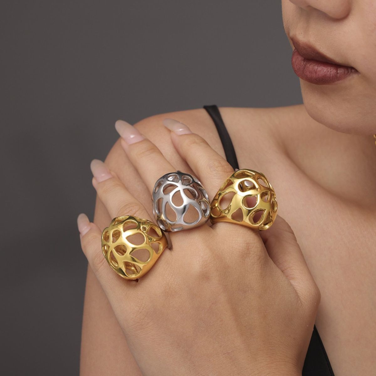 A person wearing a black strap tank top showcases three large, intricately designed rings, including the Gold-Cutout Elegance Ring from Marianela's Exclusive Shop in 18K gold-plated and silver on their fingers against a dark gray background, highlighting a touch of modern minimalism.