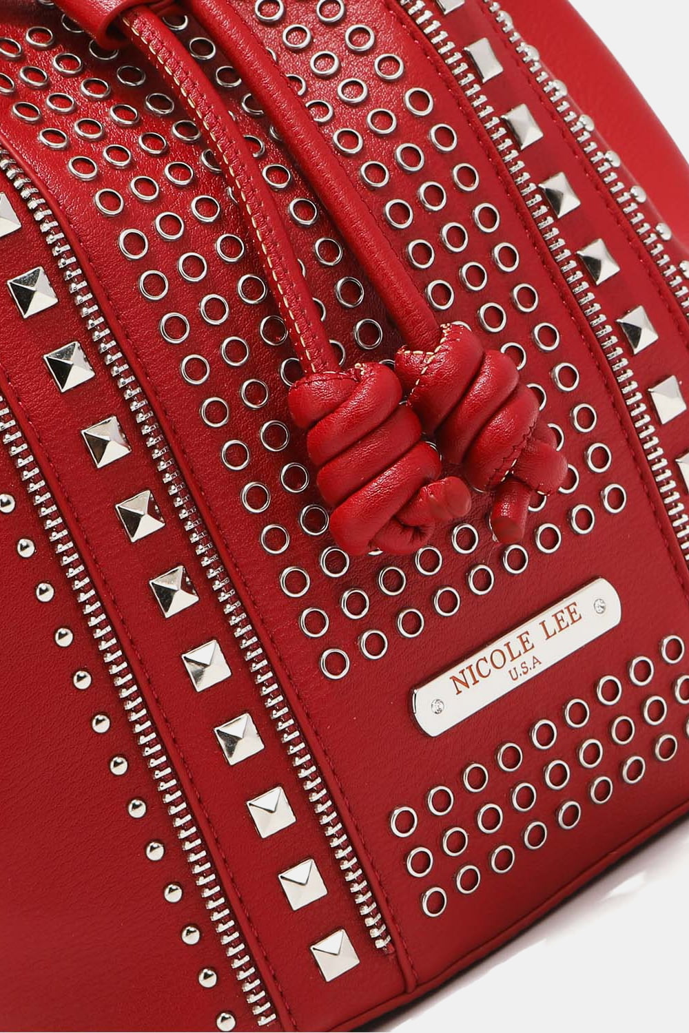 Close-up of a red Nicole Lee USA Amy Studded Bucket Bag from Marianela's Exclusive Shop, LLC, featuring intricate detailing with metal studs, circular perforations, and zippers. The bag showcases a label with the brand name "Nicole Lee USA" and has secure drawstring closure with knotted ends made from pebbled vegan leather.