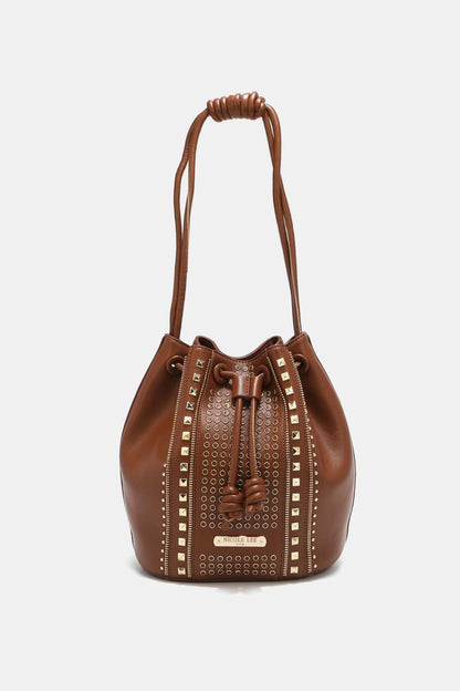 The Nicole Lee USA Amy Studded Bucket Bag from Marianela's Exclusive Shop, LLC features a brown drawstring design adorned with a pattern of gold studs and eyelets forming vertical lines on its front. It boasts a knotted handle at the top and a pebbled vegan leather finish. The bag is displayed against a plain white background.