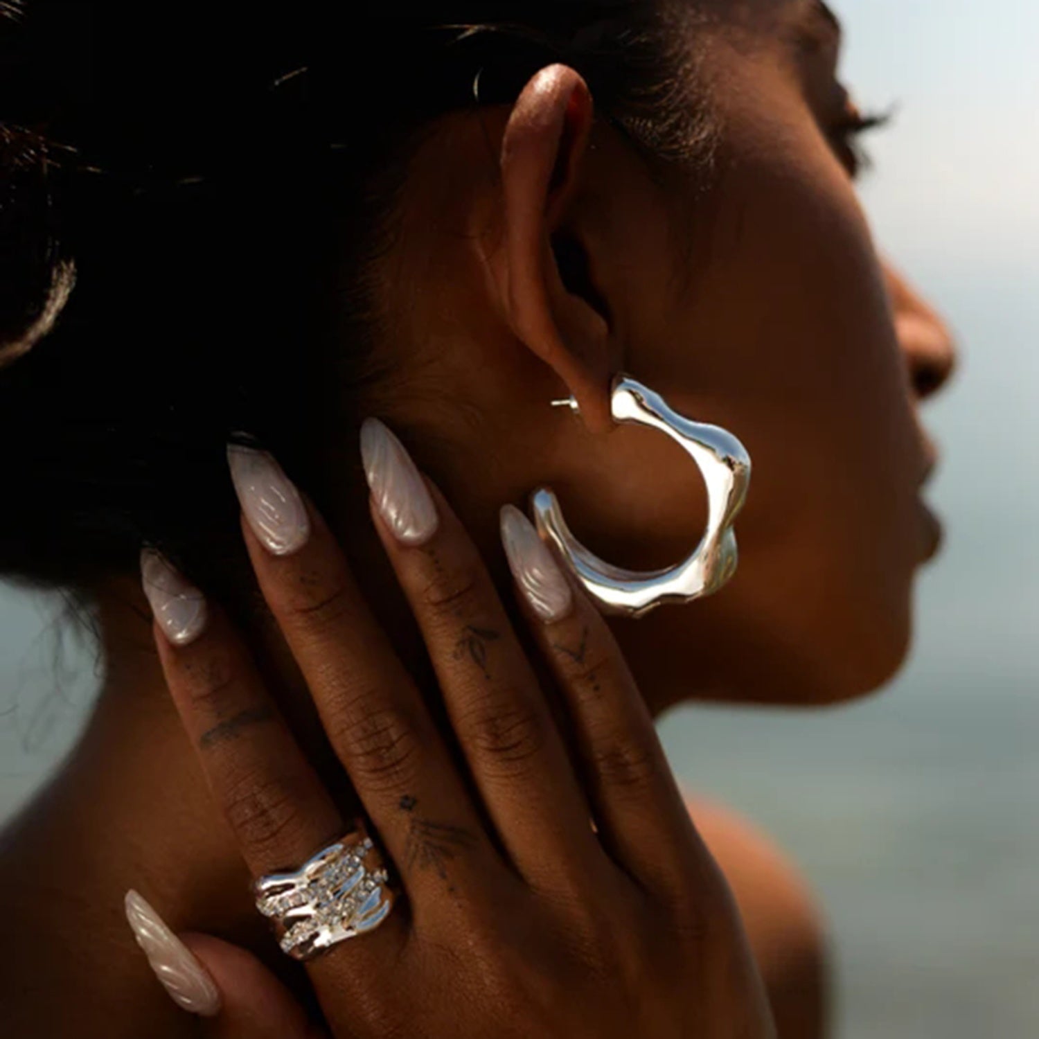 A close-up side profile of a person showcasing their sophisticated design Stainless Steel C-Hoop Earrings from Marianela's Exclusive Shop, LLC and a hand with long, manicured nails adorned with 18K gold-plated rings in focus. The background is blurred, highlighting the accessories and polished nails.