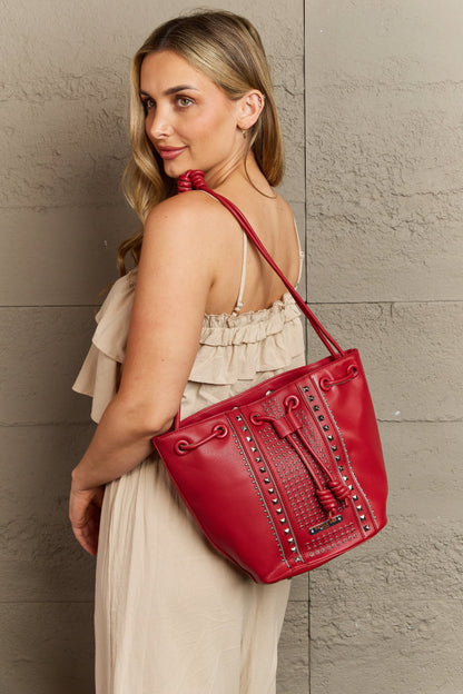 A woman with long blonde hair is gazing back over her shoulder, wearing a light beige sleeveless dress. She is carrying the Nicole Lee USA Amy Studded Bucket Bag from Marianela's Exclusive Shop, LLC, a stylish red handbag made of pebbled vegan leather with stud detailing. She stands against a textured gray wall.