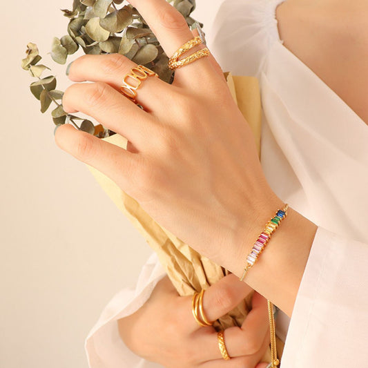 Close-up of a person’s hands adorned with various gold rings and wearing the Contrast Zircon Titanium Steel Bracelet from Marianela's Exclusive Shop, LLC. The person is holding dried eucalyptus wrapped in brown paper and is dressed in a white top with loose-fitting sleeves.