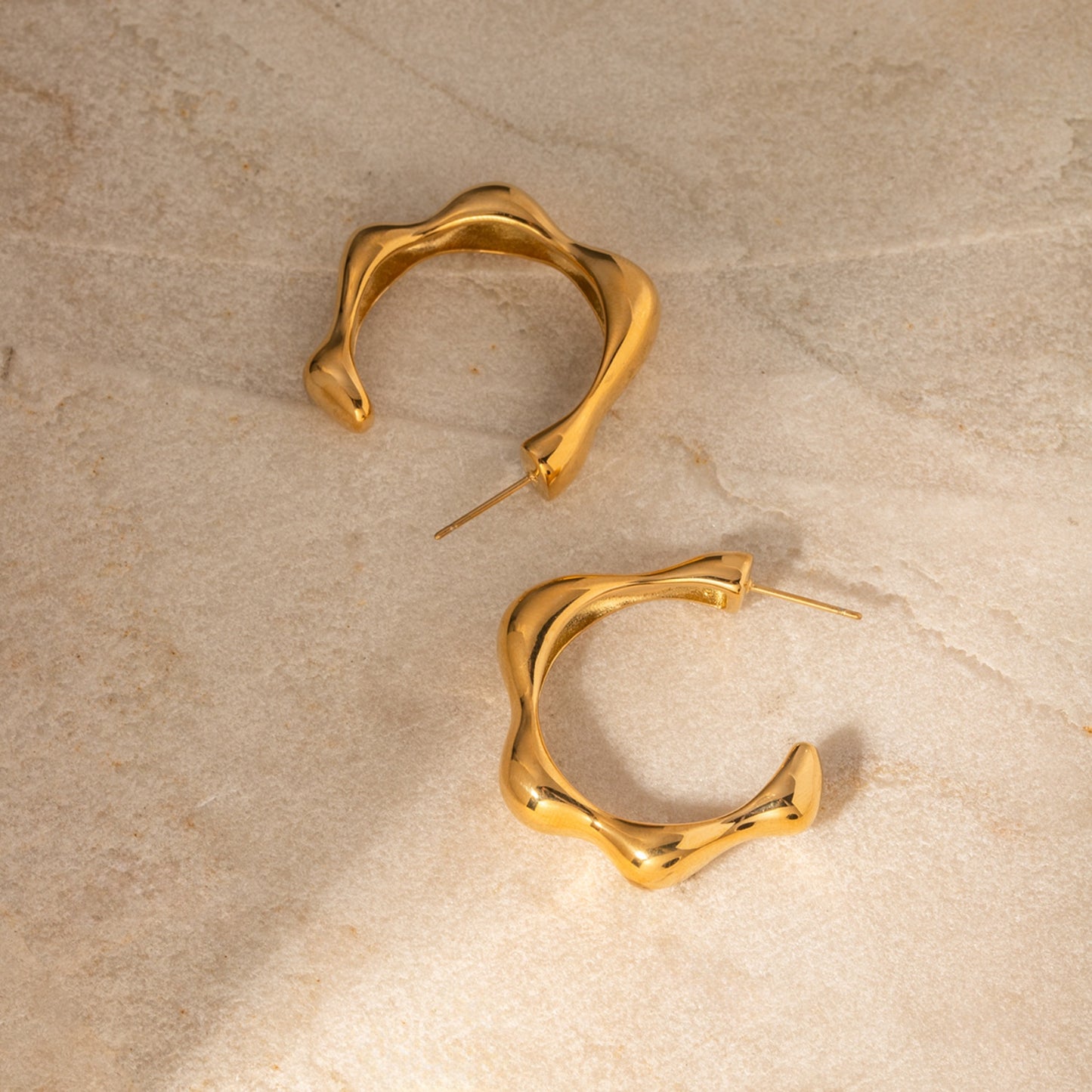 A pair of abstract, wavy stainless steel C-hoop earrings from Marianela's Exclusive Shop, LLC laid out on a warm-toned, textured stone surface. The earrings have a unique, fluid shape with a polished finish, showcasing an artistic and sophisticated design.
