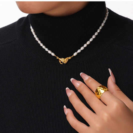 A person dressed in a black turtleneck showcases the stunning Unity Pearl Necklace from Marianela's Exclusive Shop, featuring a gold clasp elegantly around their neck. Their hand, embellished with a large gold ring, rests near their collarbone, while their nails display a pale pink polish.