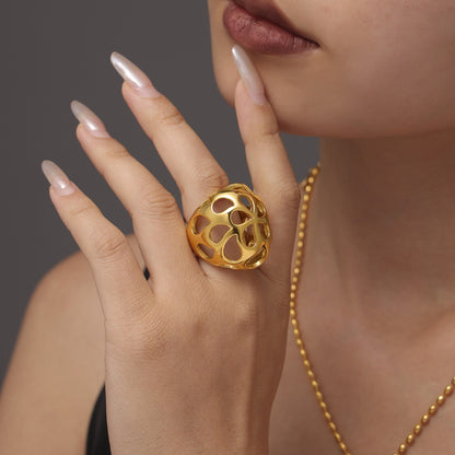 A person with nails painted in a light pink hue gently touches their face, highlighting the Gold-Cutout Elegance Ring from Marianela's Exclusive Shop, featuring a modern minimalism design in 18K gold-plated stainless steel. Complementing the look is a gold beaded necklace set against a neutral background.