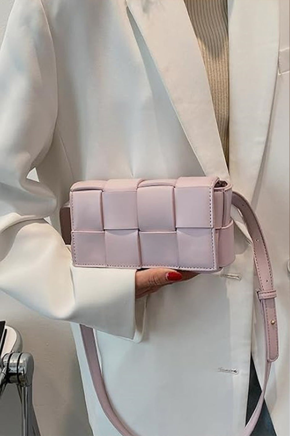 A person wearing a white blazer holds a small, pastel pink, Zenana Vegan Leather Woven Crossbody Bag from Marianela's Exclusive Shop, LLC with a long strap. The bag is detailed with a square pattern and matches the clean, sophisticated style of the blazer. The person's nails are painted red.