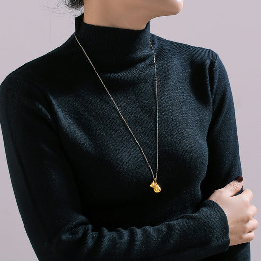 A person in a black turtleneck sweater, arms crossed, elegantly showcases the Elysian Gold Water Drop Necklace from Marianela's Exclusive Shop. The long chain features a stainless steel pendant with a water drop shape, set against a plain, light-colored background.