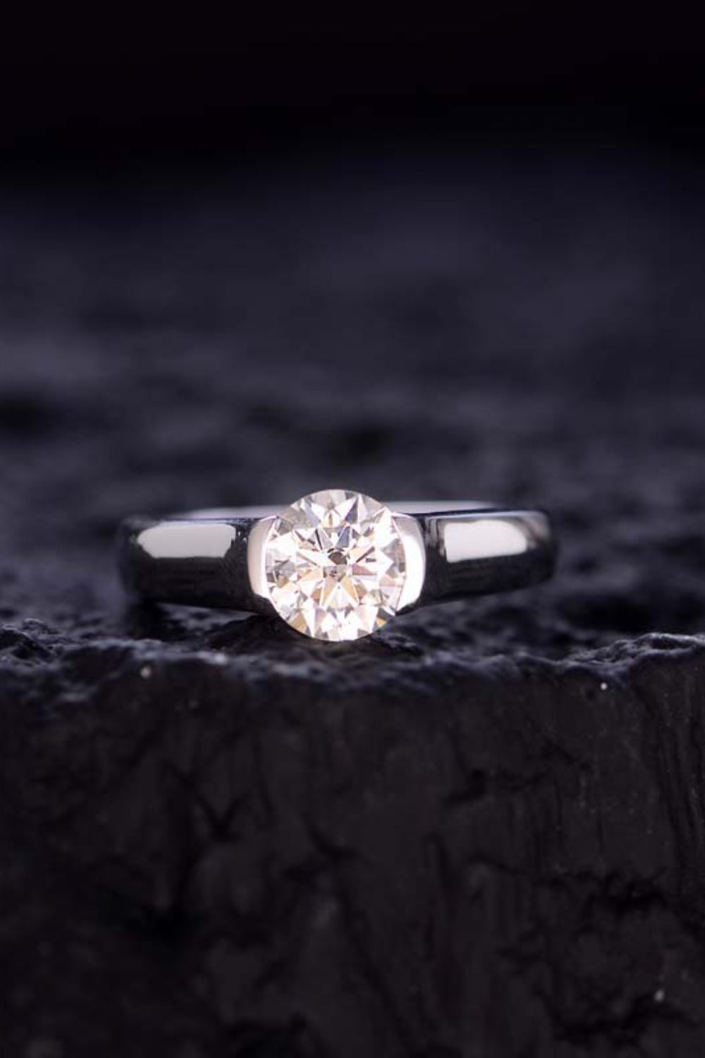 A close-up of a 1.5 Carat Moissanite 925 Sterling Silver Ring by Marianela's Exclusive Shop, LLC with a round-cut gem set on a sleek, gleaming 925 sterling silver band, placed on a textured dark background. The moissanite sparkles brightly, drawing attention to its clarity and brilliance.