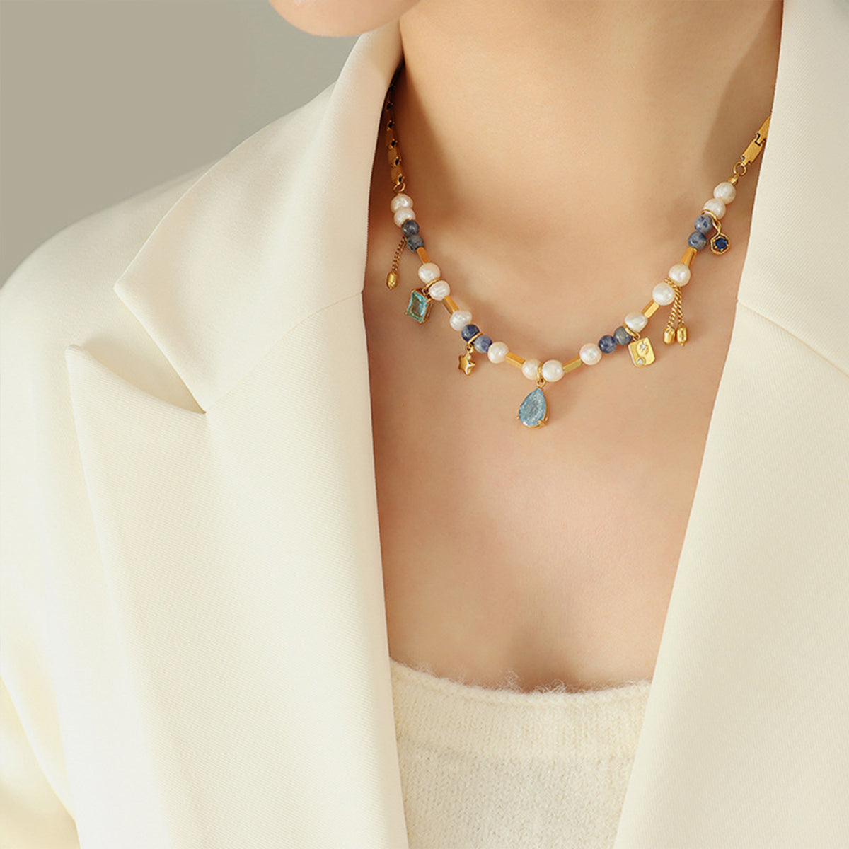 A person wearing a cream blazer and white top is adorned with Beaded Charm Necklace by Marianela's Exclusive Shop, LLC. The delicate piece features Freshwater Pearls, blue and green gemstones, and small 18K Gold-Plated charms. The person's head is not fully visible, focusing on the neckline and necklace.