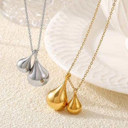 The scene showcases two exquisite necklaces from Marianela's Exclusive Shop: the Elysian Gold Water Drop Necklace, featuring dual water drop pendants in 18K gold-plated elegance, and a silver-toned stainless steel piece with three teardrop-shaped pendants. Both are artfully displayed on a textured, light-hued background adjacent to a white patterned plate.