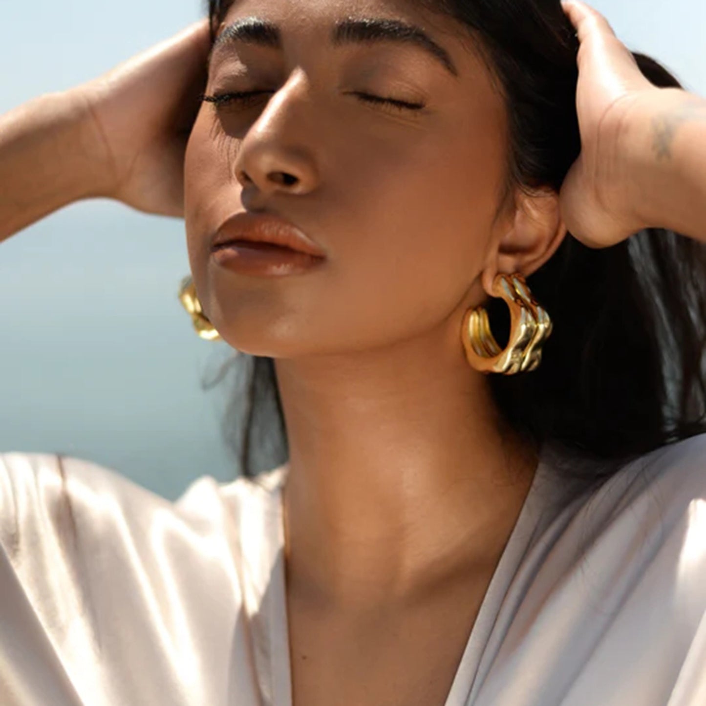 A woman with dark hair and closed eyes tilts her head back, hands touching her head. She wears large, sophisticated Stainless Steel C-Hoop Earrings from Marianela's Exclusive Shop, LLC and a silky light-colored top. The background is blurred, suggesting an outdoor setting, possibly near water.
