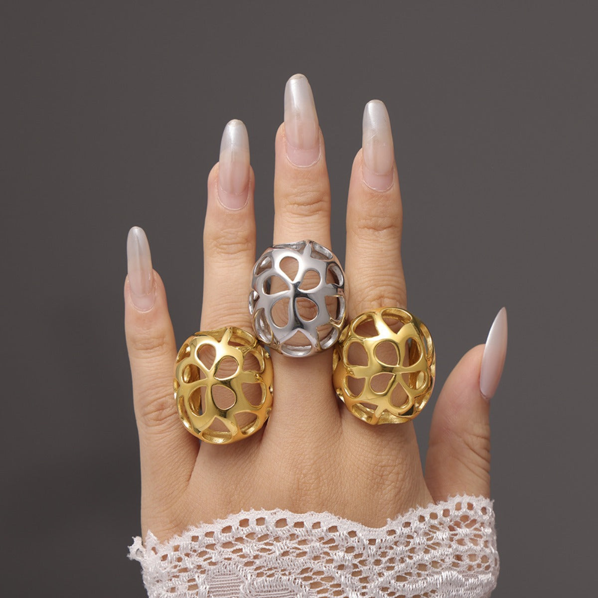 A hand with lace accentuating the wrist showcases three large, ornate rings. Both outer rings are 18K gold-plated, while the center one is a Stainless Steel Gold-Cutout Elegance Ring from Marianela's Exclusive Shop. These rings feature an intricate floral, openwork design. The nails are long and polished in a pearly hue, reflecting modern minimalism.
