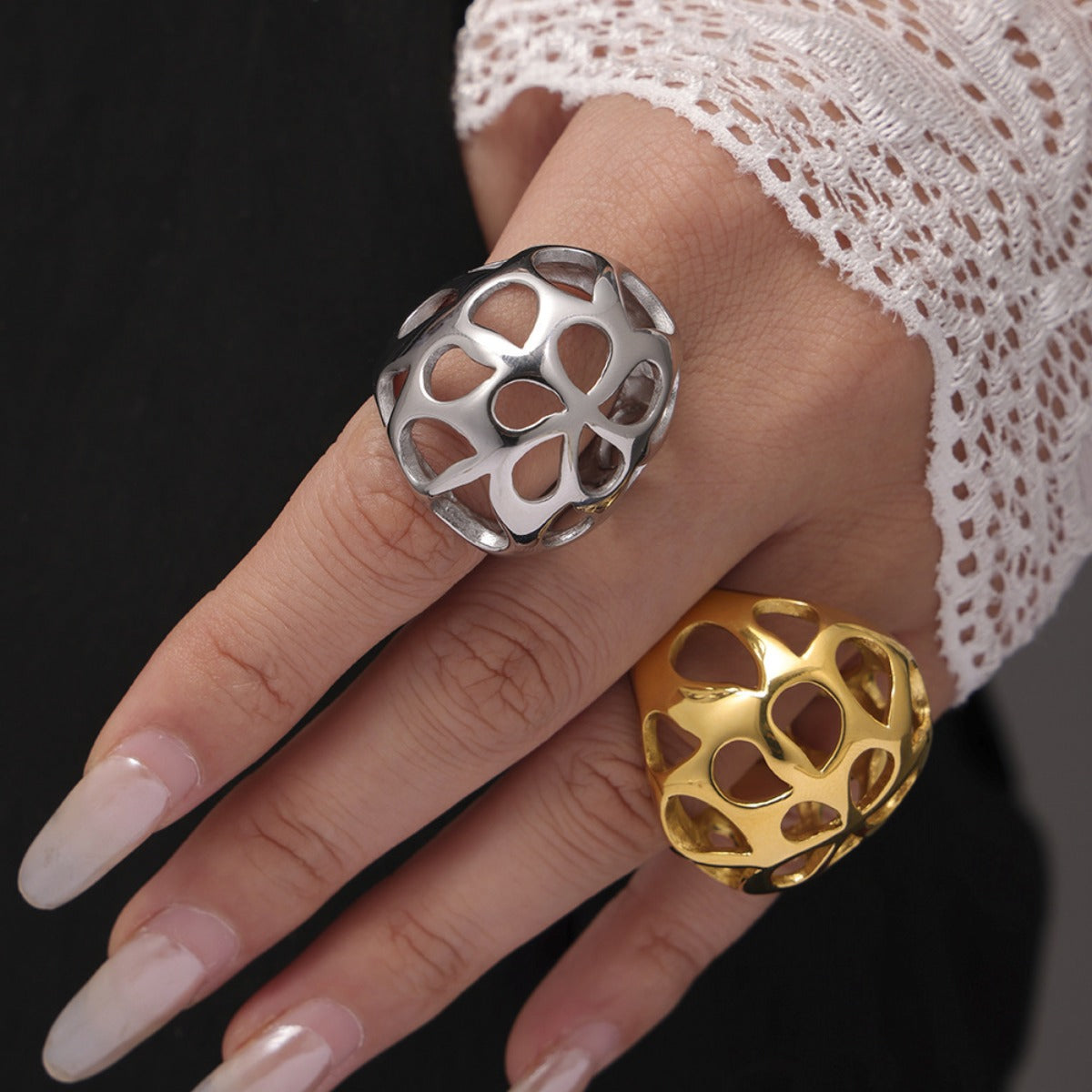 A hand with long, manicured nails and a lace sleeve showcases two statement rings: the Gold-Cutout Elegance Ring from Marianela's Exclusive Shop, plated in 18K gold, and a Stainless Steel Cutout Ring. Both feature heart-shaped designs on the middle and ring fingers, embodying modern minimalism against a dark background.