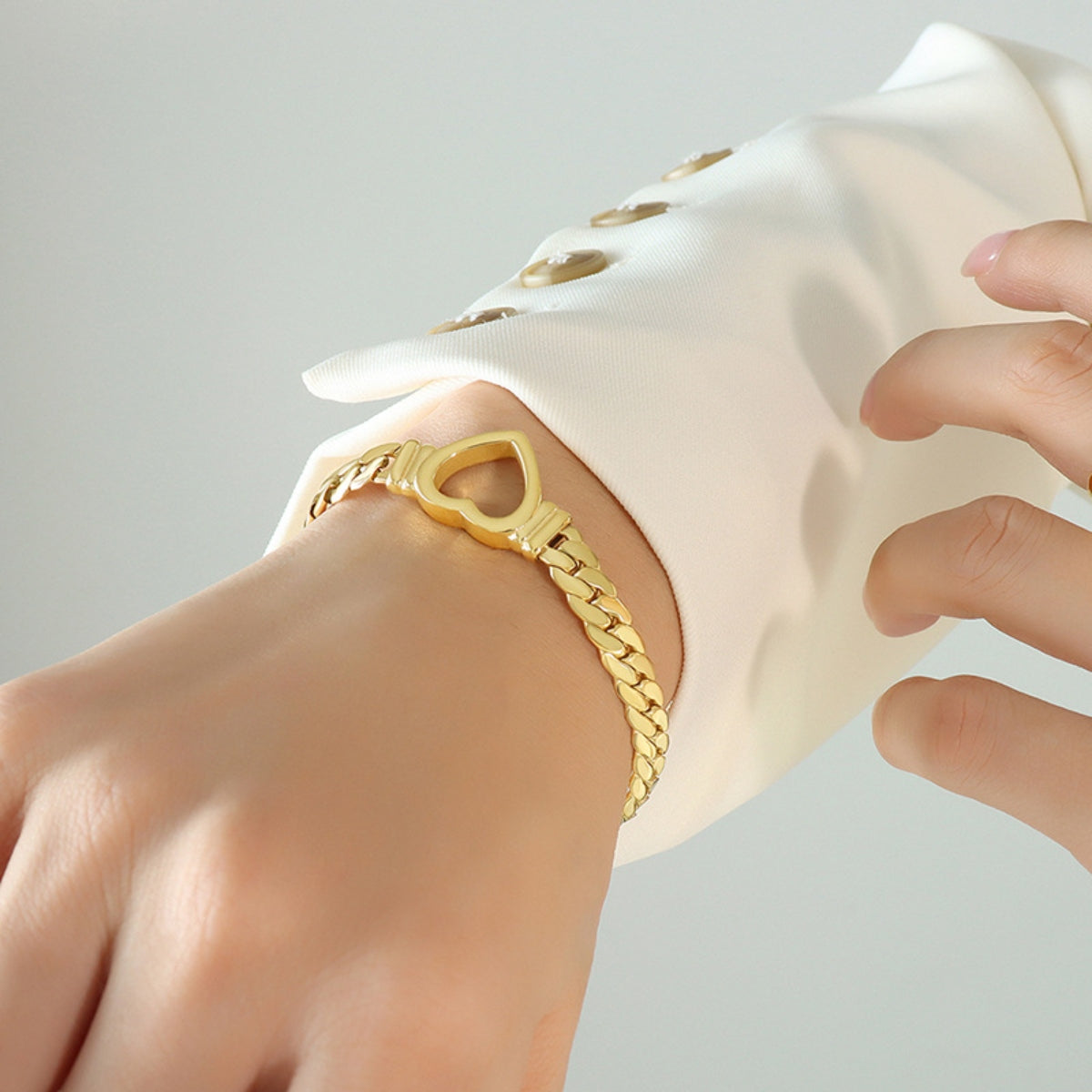 A close-up image of a person's wrist adorned with the Titanium Steel Cutout Heart Bracelet from Marianela's Exclusive Shop, LLC. The person is wearing a white long-sleeved shirt with beige buttons, which can be seen in the background.