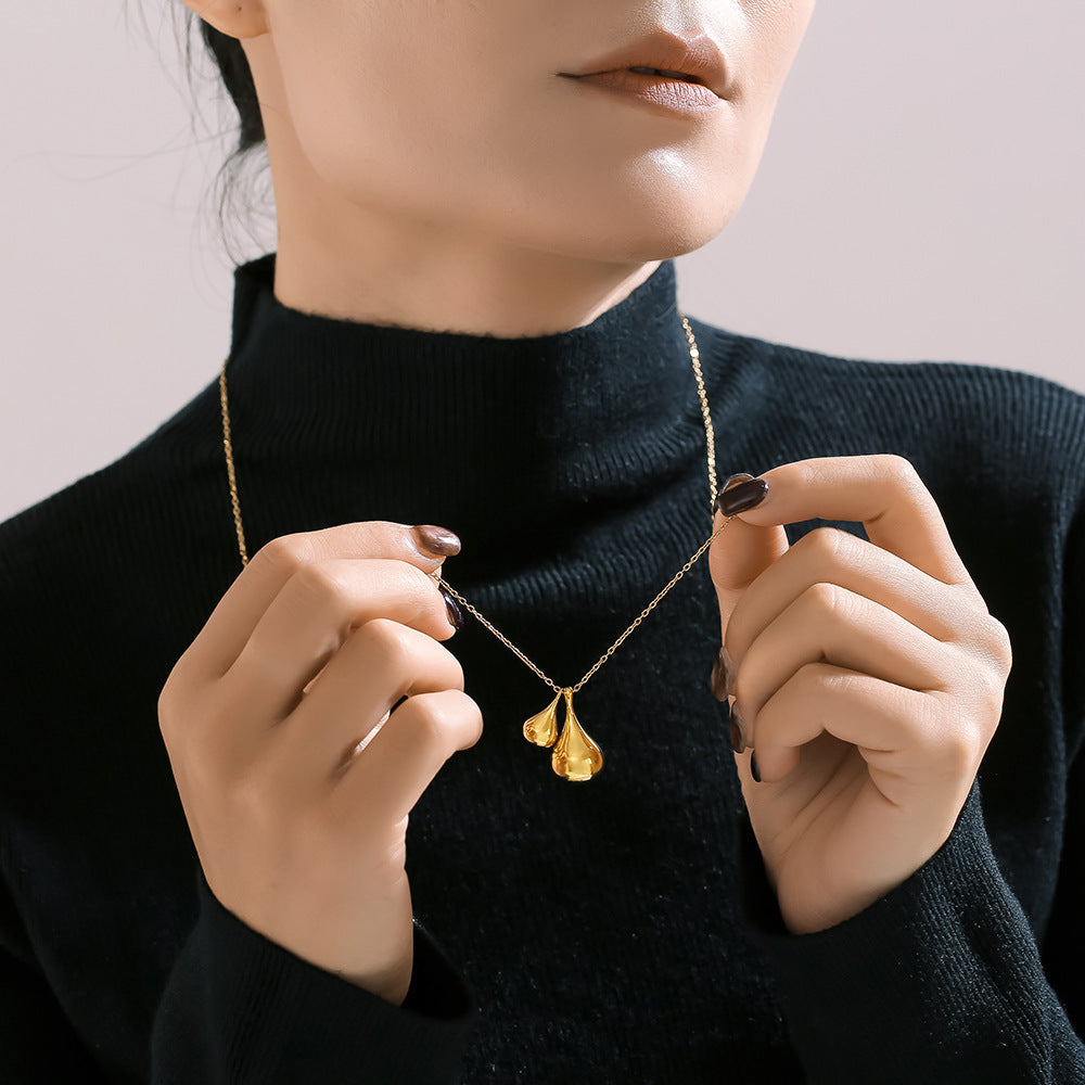 An individual dressed in a black turtleneck sweater is holding an Elysian Gold Water Drop Necklace by Marianela's Exclusive Shop, featuring an 18K gold-plated pendant shaped like two water drops. Their nails are painted dark brown and they wear a neutral lip shade, while the softly lit background accentuates the necklace's elegance.