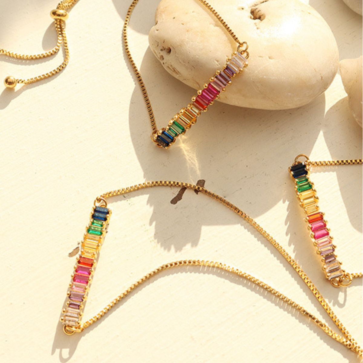 Gold chains adorned with rectangular, multicolored gemstone pendants are displayed against a light-colored background with smooth white stones. The pendants feature a mix of vibrant pink, green, blue, red, and clear gems, highlighting their intricate design alongside the Contrast Zircon Titanium Steel Bracelet from Marianela's Exclusive Shop, LLC.