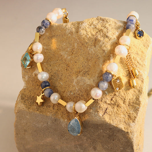 A Beaded Charm Necklace with a mix of blue and white beads, gold accents, and a teardrop-shaped blue pendant is draped over a rough, beige rock. The 18K gold-plated necklace from Marianela's Exclusive Shop, LLC also features small star and hexagon charms, freshwater pearls, and additional gold and blue decorative elements.