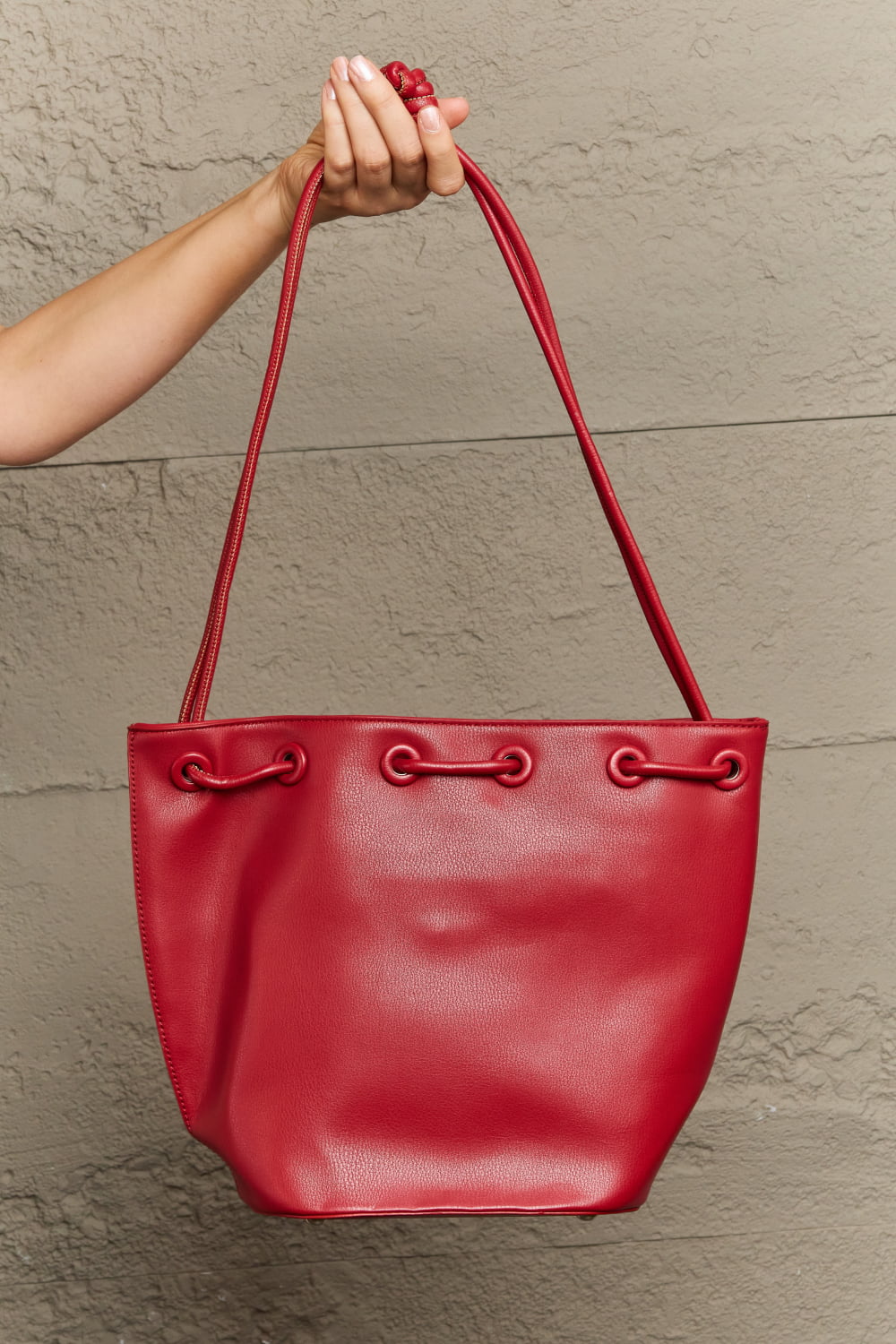 A person holding the chic Nicole Lee USA Amy Studded Bucket Bag, a stylish red tote from Marianela's Exclusive Shop, LLC. This bag, crafted from pebbled vegan leather, boasts two long handles and a secure drawstring closure with looped details near the top. Subtle stud detailing completes its elegant look against a textured gray wall backdrop.