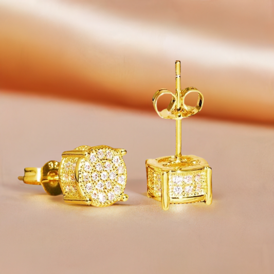 A pair of trendy Bling Studs Gold earrings from Marianela's Exclusive Shop, LLC adorned with shiny zircons. One earring is lying on its side, showcasing the clustered zircon pattern on the stud, while the other stands upright, highlighting the earring post and butterfly back. Perfect for daily wear, they're set against a soft, muted peach background.