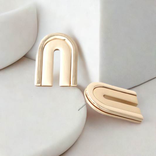 A pair of gold-colored, double-layer **Arch Earrings Studs** with a sleek, modern design. Placed on a soft white surface, they create a minimalist and elegant aesthetic. The lighting highlights their smooth texture and polished finish, perfect for any daring fashion icon seeking versatility. From **Marianela's Exclusive Shop**.