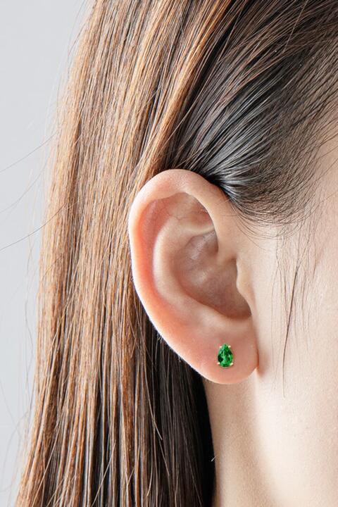 Close-up of a woman's ear adorned with Marianela's Exclusive Shop, LLC Emerald Stud Earrings featuring a small lab-grown emerald set in 925 sterling silver. Her straight brown hair is tucked behind her ear, and the background is a plain, soft gray.