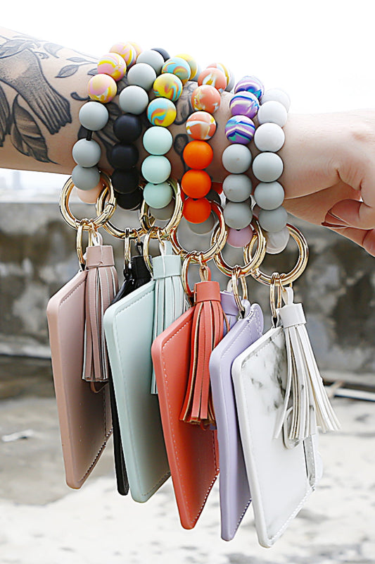 A person's tattooed arm holds several wristlet keychains with Marianela's Exclusive Shop, LLC Beaded Tassel Keychain with Wallet. The bracelets feature various colorful bead patterns, while the durable leather cardholders come in different colors and materials. The background shows an outdoor setting.