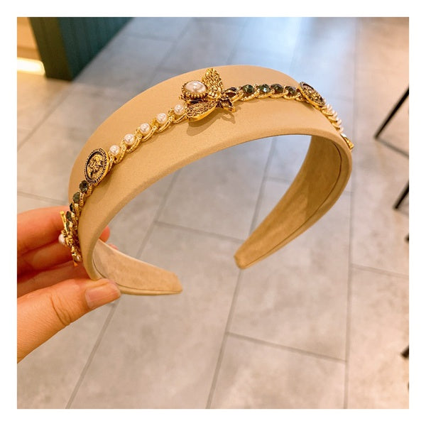 A beige hand-crafted headband adorned with a row of pearls, gold buttons, and intricate gold flower and leaf embellishments is held by a hand against a tiled floor background, showcasing the elegance of Baroque Glam Rhinestone Headbands by Marianela's Exclusive Shop, LLC.