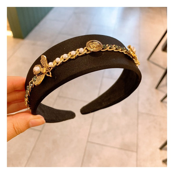 A black headband adorned with decorative gold embellishments, including a bee charm, pearl-like beads, and a gold chain, is held by a hand against a blurred background featuring tiled flooring and partially visible furniture. This Baroque Glam Rhinestone Headbands from Marianela's Exclusive Shop, LLC perfectly showcases the elegance of rhinestones and metals.