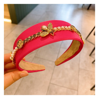 A hand holds a vibrant pink, hand-crafted headband adorned with decorative elements. The Baroque Glam Rhinestone Headbands from Marianela's Exclusive Shop, LLC features rhinestone and pearl accents, including a prominent gold bee with a pearl body, and a sequence of small gold coins and beads along the top.