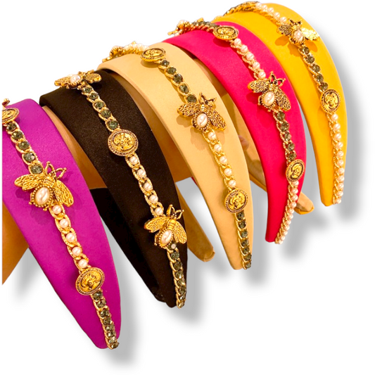 Six colorful hand-crafted headbands, from left to right: purple, black, yellow, hot pink, beige, and yellow. Each headband is adorned with gold-toned accents such as bees, pearls, and coins in a decorative pattern. These Baroque Glam Rhinestone Headbands from Marianela's Exclusive Shop, LLC are arranged in a row.