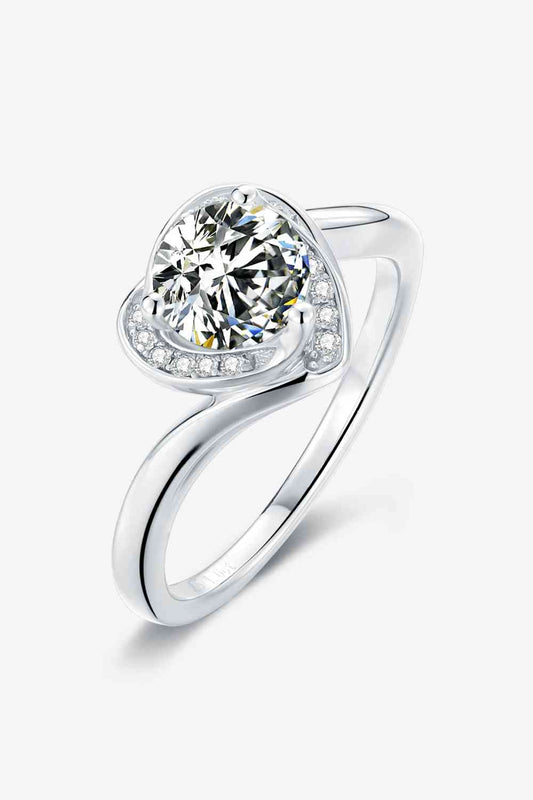 A 1 Carat Moissanite 925 Sterling Silver Heart Ring by Marianela's Exclusive Shop, LLC showcases a large round-cut diamond at the center, flanked by a halo of smaller diamonds. The band elegantly twists to emphasize the central gemstone, providing a modern and sophisticated design with a minimalist style. The background is white.