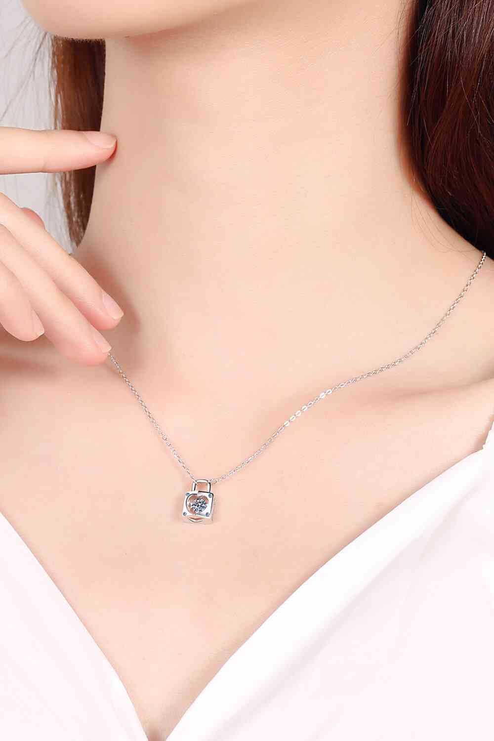 A person wearing a delicate 925 sterling silver necklace with a small square pendant featuring a single moissanite gem in the center. The Adored Moissanite Lock Pendant Necklace from Marianela's Exclusive Shop, LLC is prominently displayed. The person is dressed in a white top, and their hand is gently touching their chin. The background is light, highlighting the jewelry beautifully.